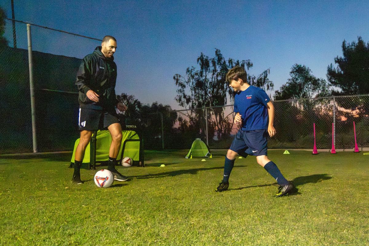 A player dribbles past a grinning Coach Luka at dusk, The Soccer Lab, Carlsbad