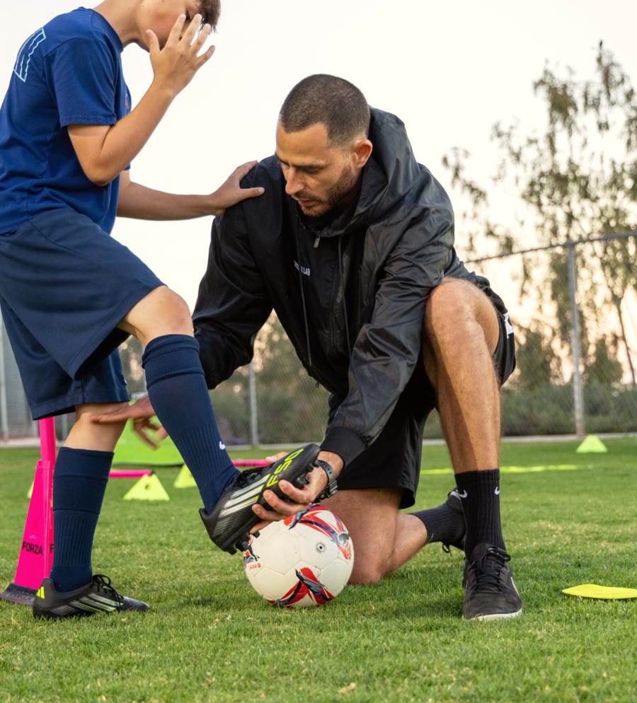 A young player steadies himself on Coach Luka's shoulder while Luka sets his foot on the ball, The Soccer Lab, Carlsbad