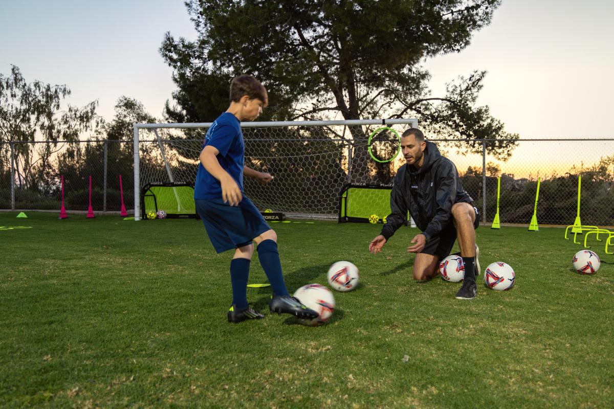 Coach Luka kneeling at eye level, watching a young player's touch at dusk, The Soccer Lab, Carlsbad